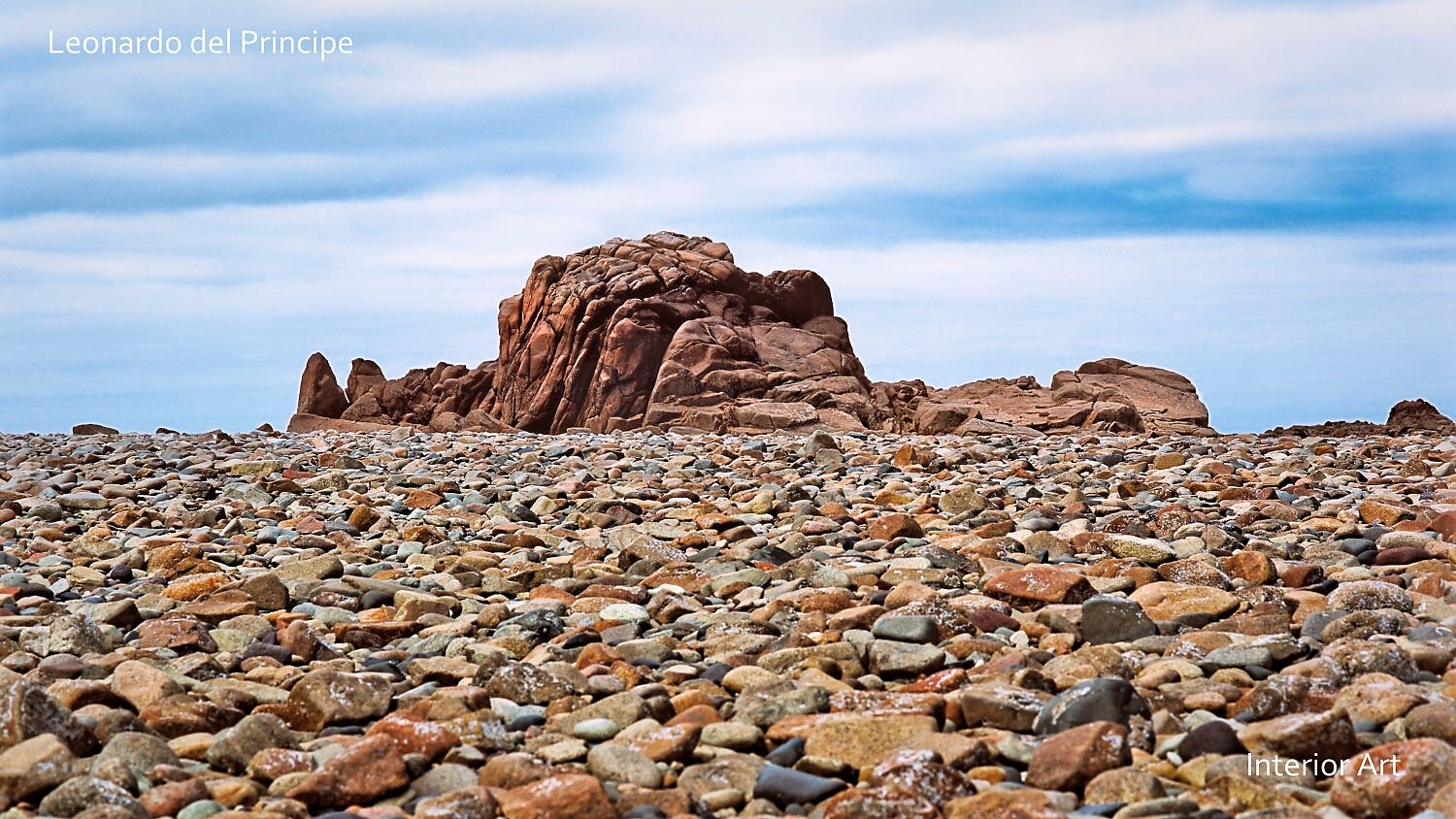MORG01 Rocky landscape with large, rugged red-brown rocks under a cloudy blue sky. Foreground is covered with smooth stones, conveying a monumental natural scene.