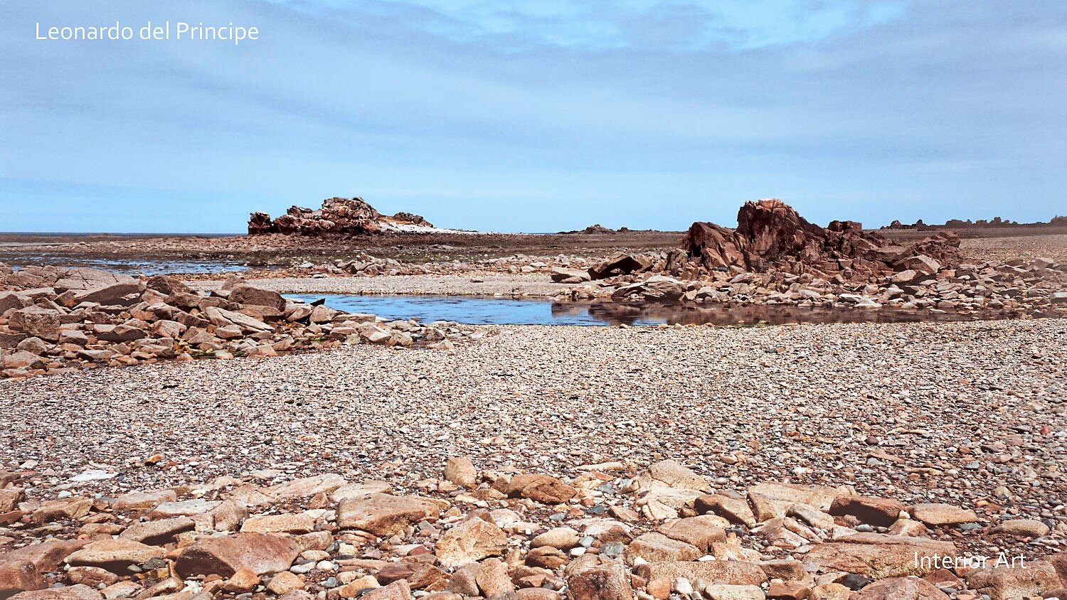 MORG02 Rocky shoreline with scattered boulders under a blue sky, distant jagged rocks near a calm, reflective tidal pool. A natural coastal scene.