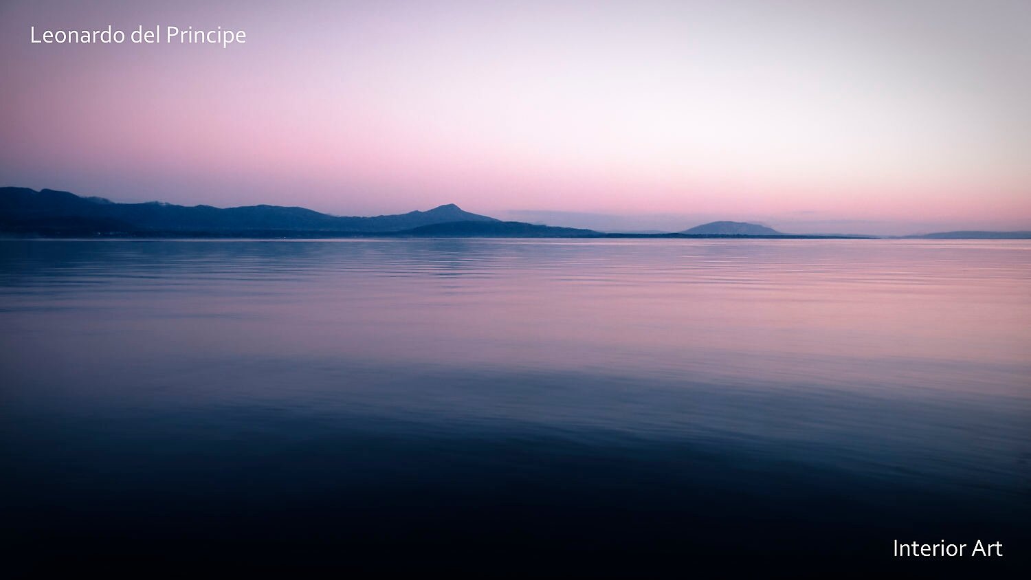 WFNF04 Calm lake at dawn with serene pink and blue hues reflecting on the water. Distant mountains line the horizon under a smooth, pastel sky.